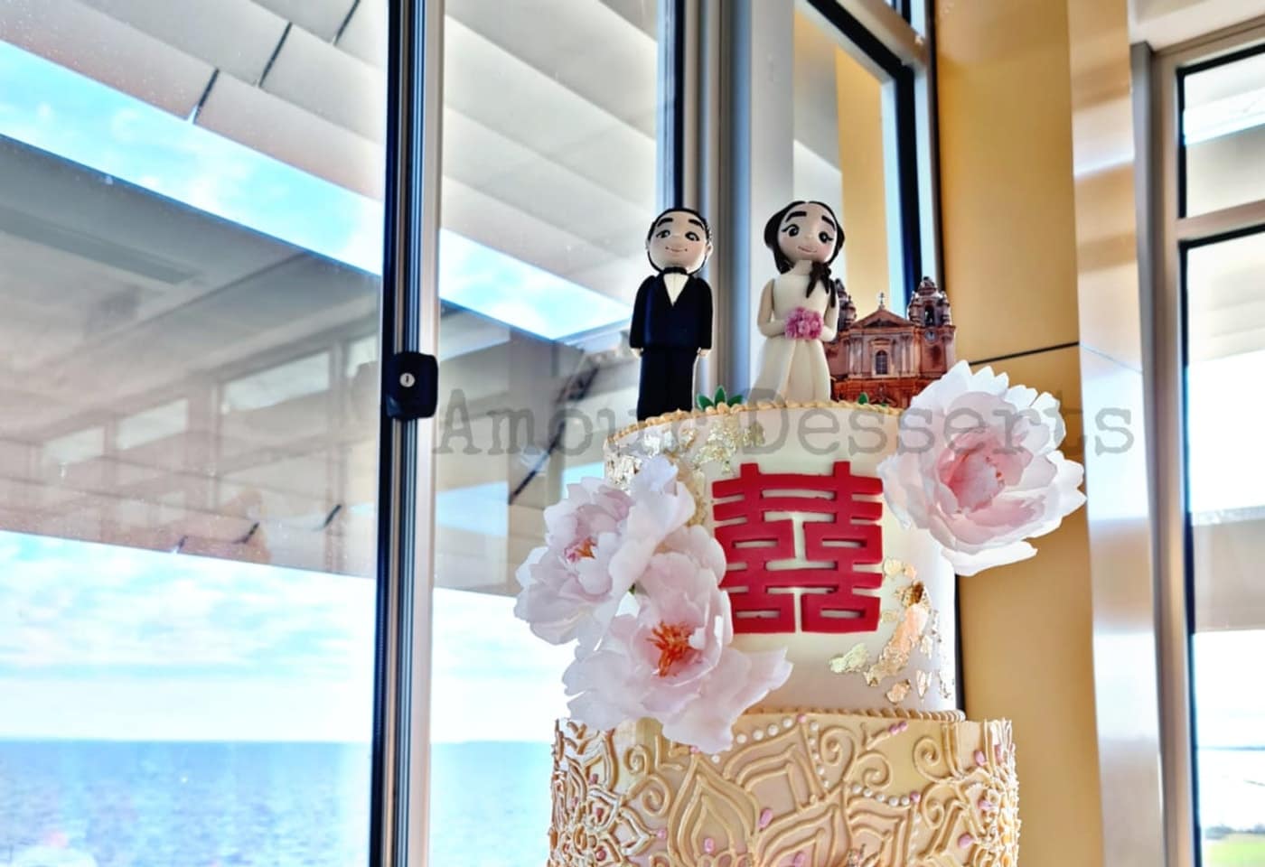 Gold lace custom cake with pink peonies, a double happiness symbol, and hand-made couple toppers by a bayside window